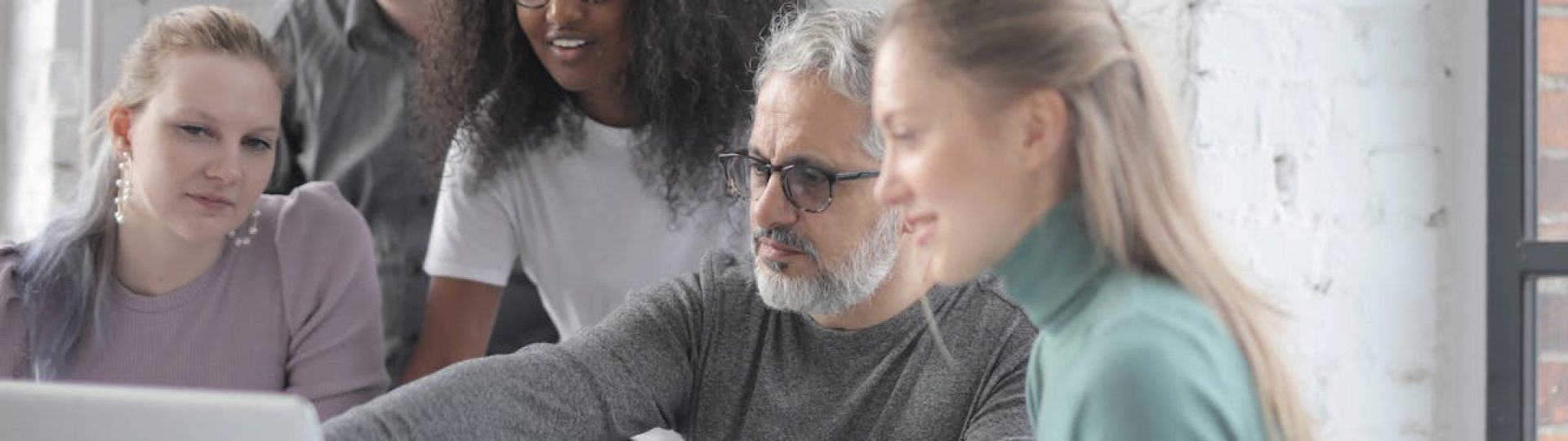 Group of diverse colleagues sitting and standing near table with laptop while discussing project and working together during business meeting