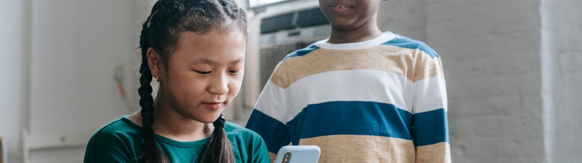 Happy little African American boy standing near Asian classmate girl using smartphone sitting at desk during break
