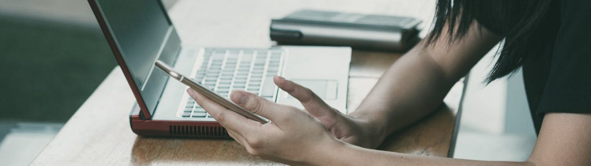 A young woman multitasking with her smartphone and laptop at a table indoors.