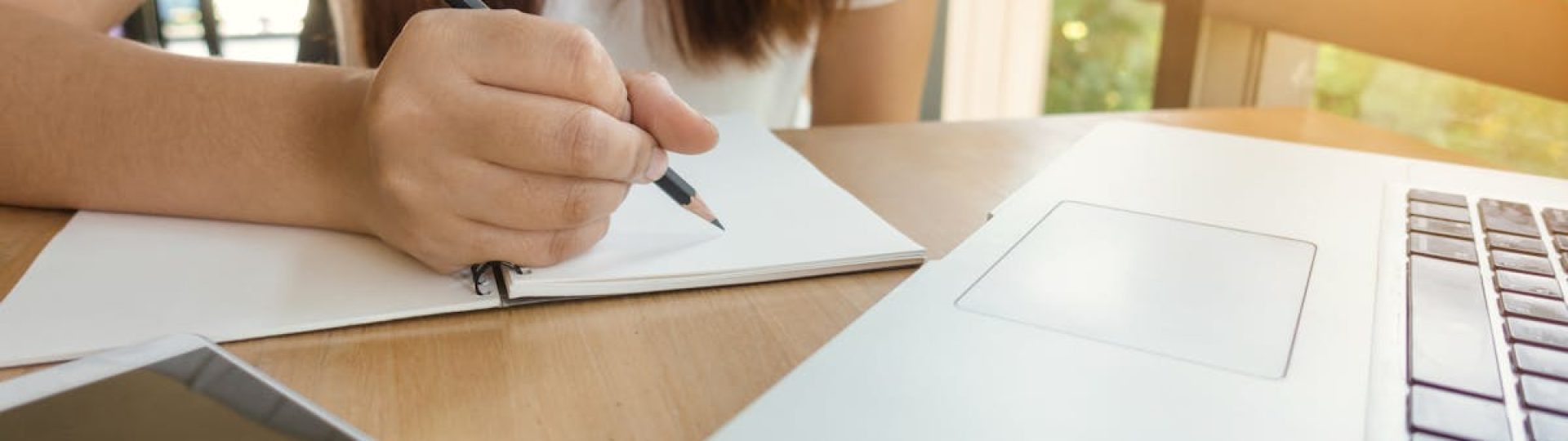 A young woman writing notes at a table with laptop and smartphone.