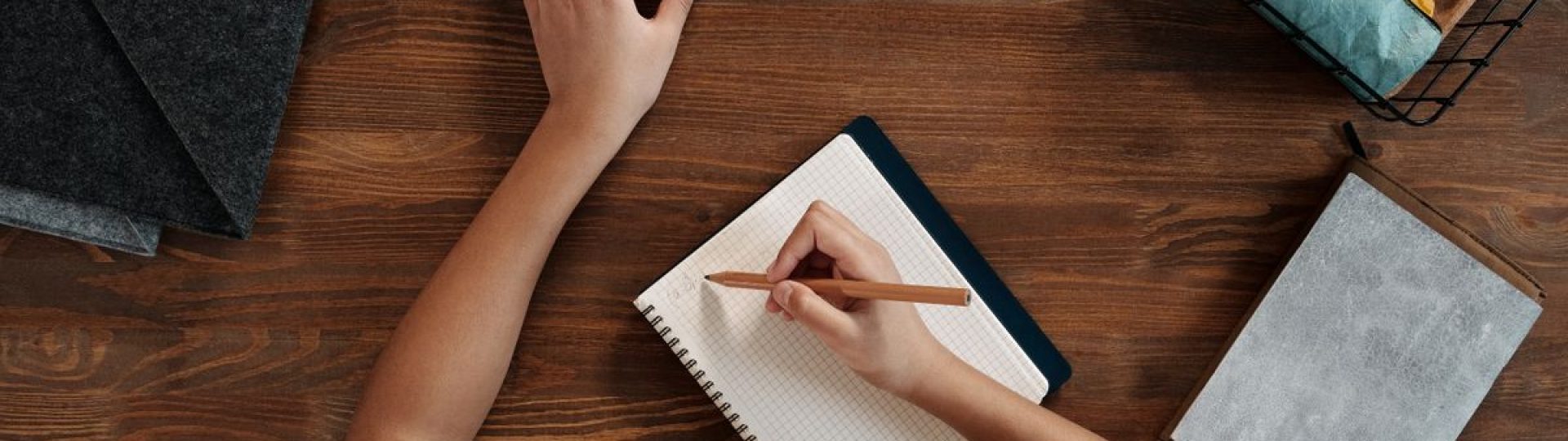 Overhead view of a child writing in a notebook while using a smartphone at a wooden desk.