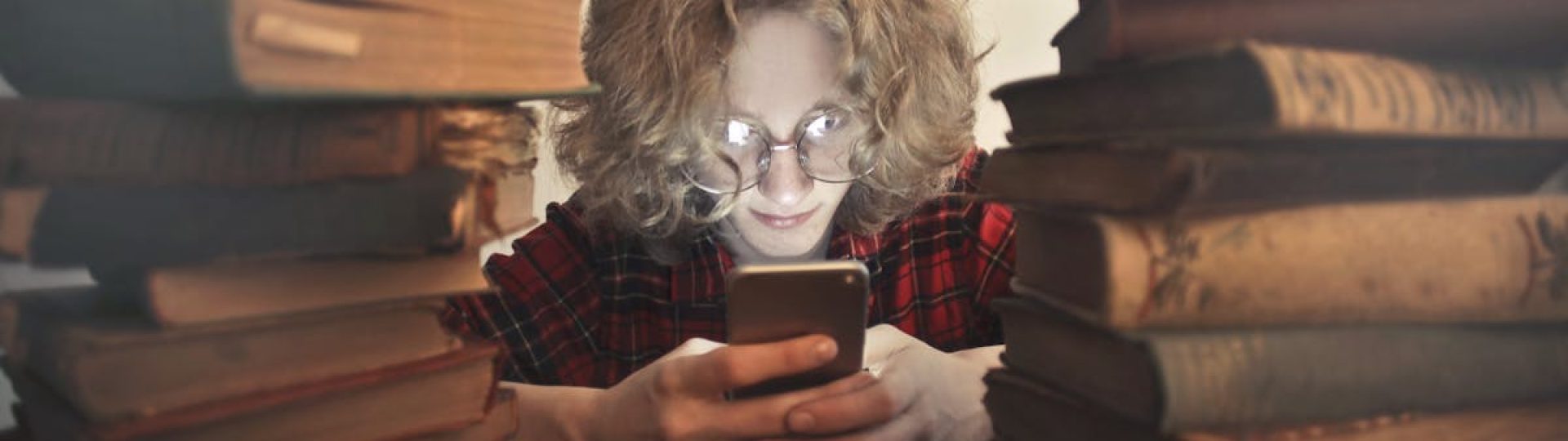 Focused male student in casual wear and glasses browsing smartphone while sitting at wooden table with stacked old books
