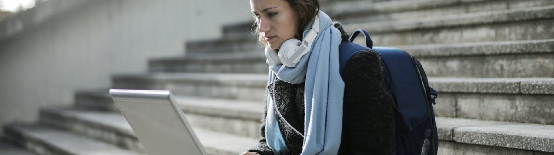 Woman using laptop on outdoor stairs, studying online with headphones and backpack.