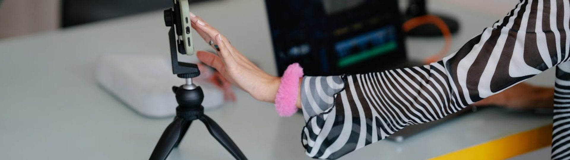 Woman adjusting smartphone on tripod with laptop setup for video editing.